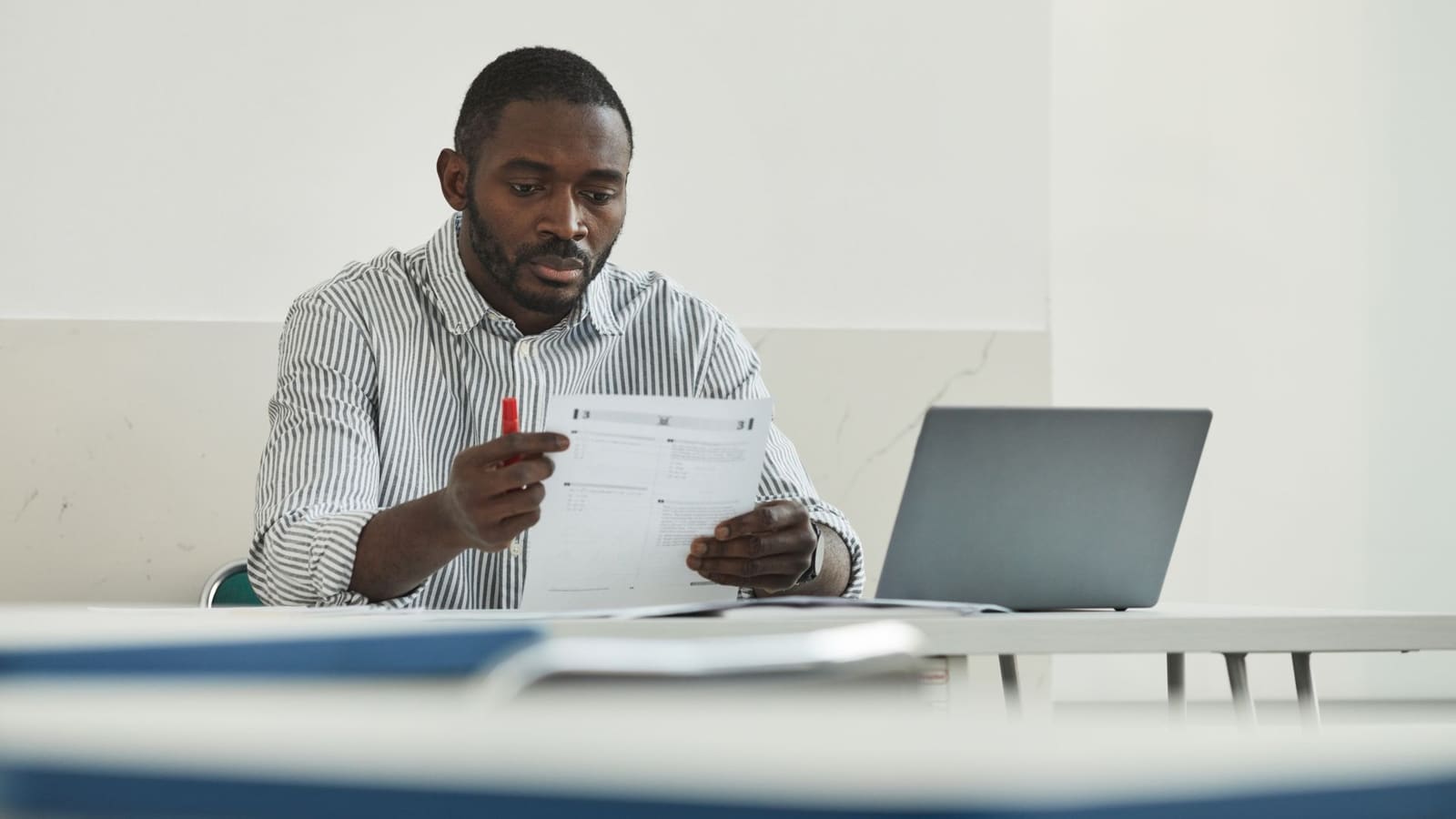 A teacher checking exam papers by hand with a laptop open beside them